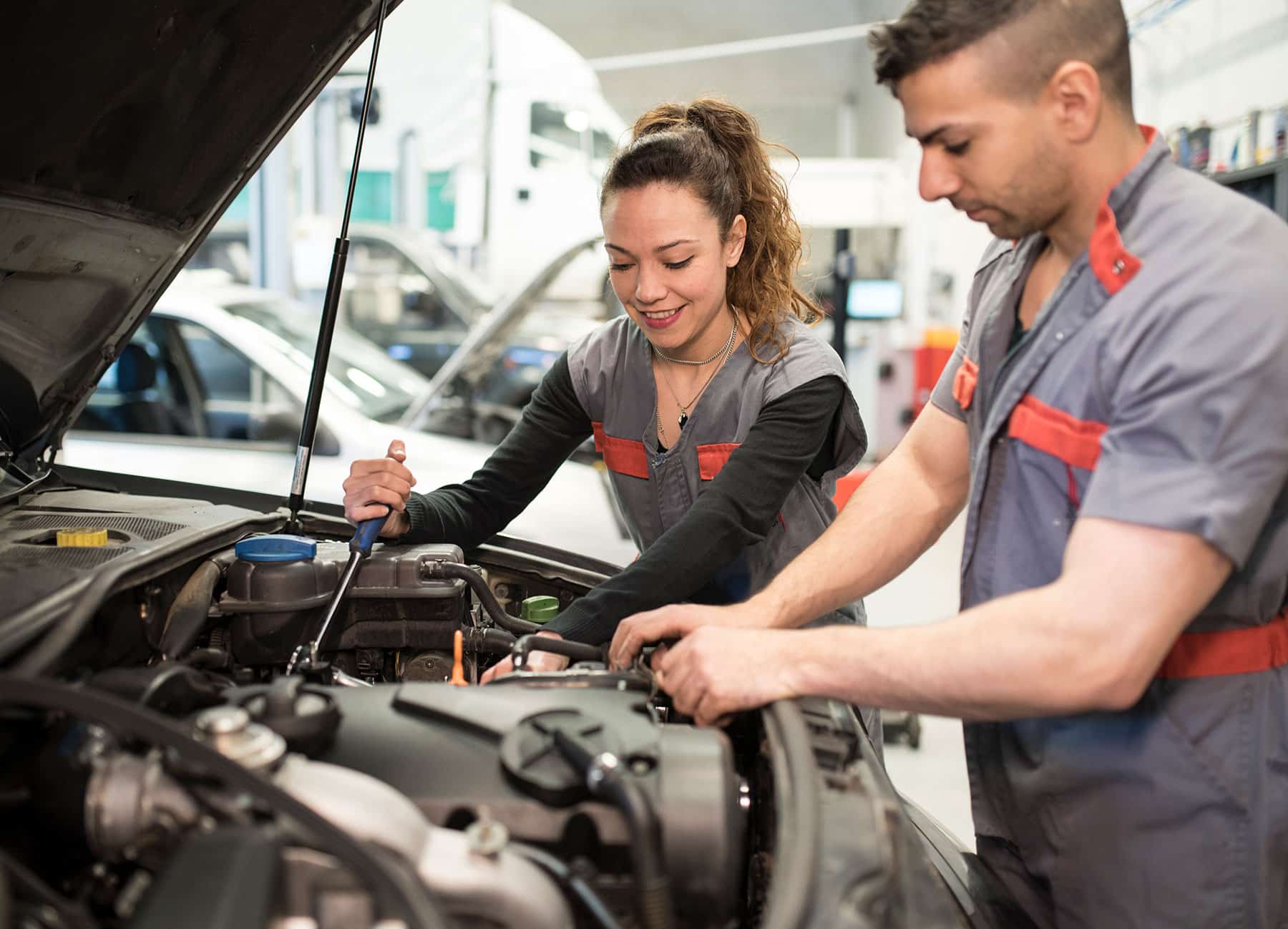 Two mechanics in gray and red uniforms work under an open car hood, inspecting the engine together in a workshop.